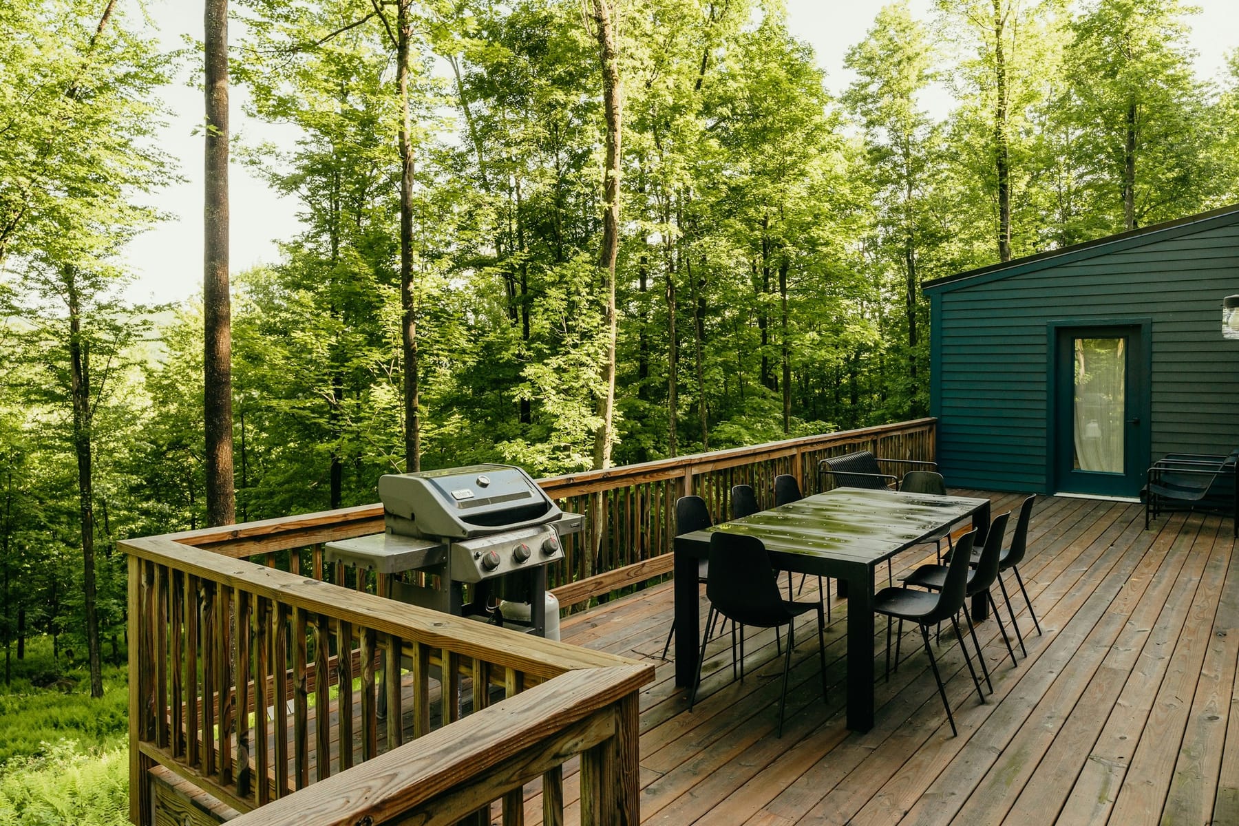 A wooden deck with a grill and a long dining table, backing straight onto mature woods.