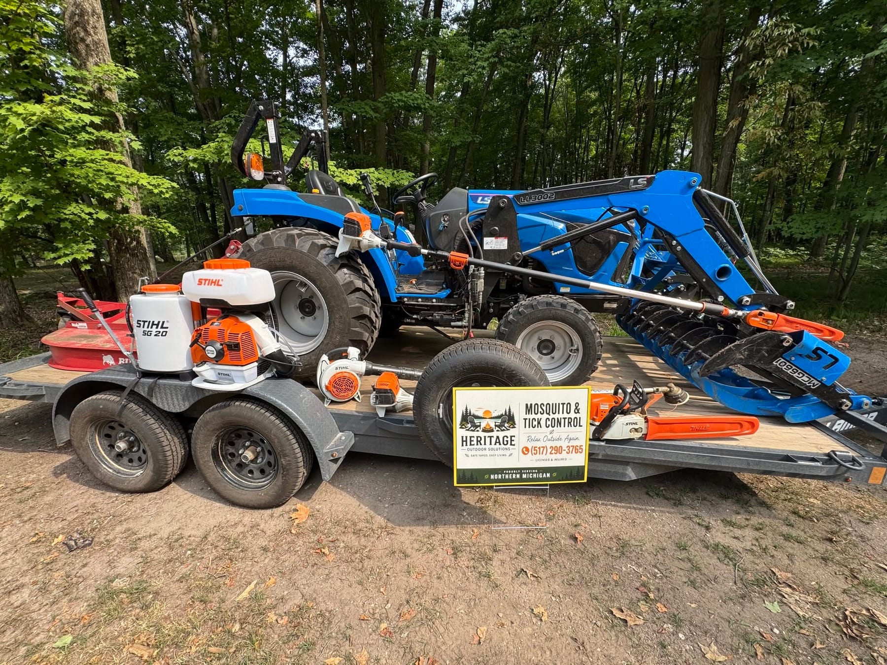 Heritage's tractor on a trailer, a hydraulic grapple mounted on its front loader and a red rotary cutter stowed beside it, with two STIHL backpack sprayers and a chainsaw on the deck and the company's yard sign propped against the trailer.